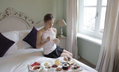 Calm female in elegant outfit sitting on bed and eating delicious breakfast served on tray in hotel room with classic interior in morning