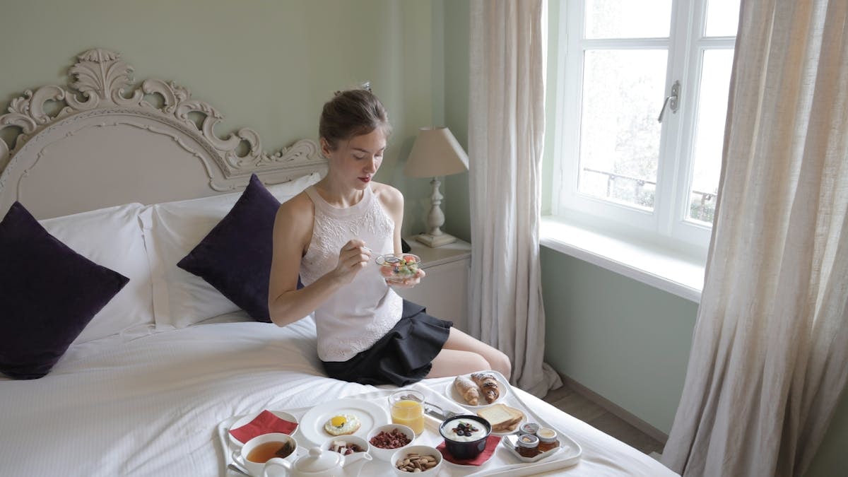 Calm female in elegant outfit sitting on bed and eating delicious breakfast served on tray in hotel room with classic interior in morning